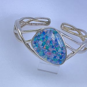 May include: Silver-toned cuff bracelet with an open design. The bracelet features a large, triangular-shaped centerpiece filled with a mosaic of blue, green, and purple iridescent pieces. The bracelet is displayed on a clear stand.