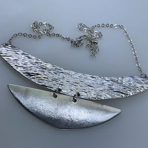 May include: A silver necklace with a textured, crescent-shaped pendant and a smaller, smooth, crescent-shaped pendant hanging below it.