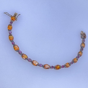 May include: A delicate bracelet featuring oval-cut orange gemstones interspersed with smaller, round, purple gemstones. The bracelet has a gold-colored clasp and is set against a light background. This piece of jewelry is suitable for special occasions.