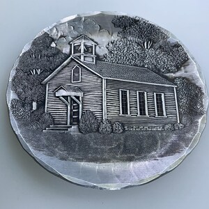 May include: A silver metal plate with a raised design of a small, wooden church with a bell tower. The church is surrounded by trees and bushes.