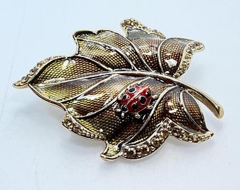 May include: A gold-tone brooch in the shape of a leaf with a red ladybug on it. The leaf is decorated with a textured pattern and rhinestones.