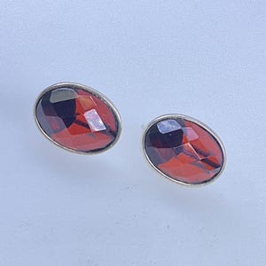 May include: A pair of oval-shaped stud earrings. Each earring features a faceted, deep red gemstone set in a silver-colored bezel. The gemstones have a mix of dark and light red facets.