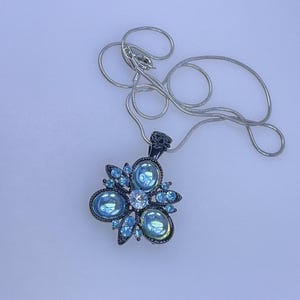 May include: A silver-toned necklace with a pendant featuring a cluster of light blue oval and round gemstones. The pendant has a dark metal setting and a delicate, silver chain. The gemstones are arranged in a floral or starburst pattern.