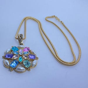 May include: A gold-toned necklace with a pendant featuring a floral design. The pendant is adorned with colorful gemstones in shades of blue, pink, and purple, surrounded by faux pearls. The chain is a thin, textured gold.