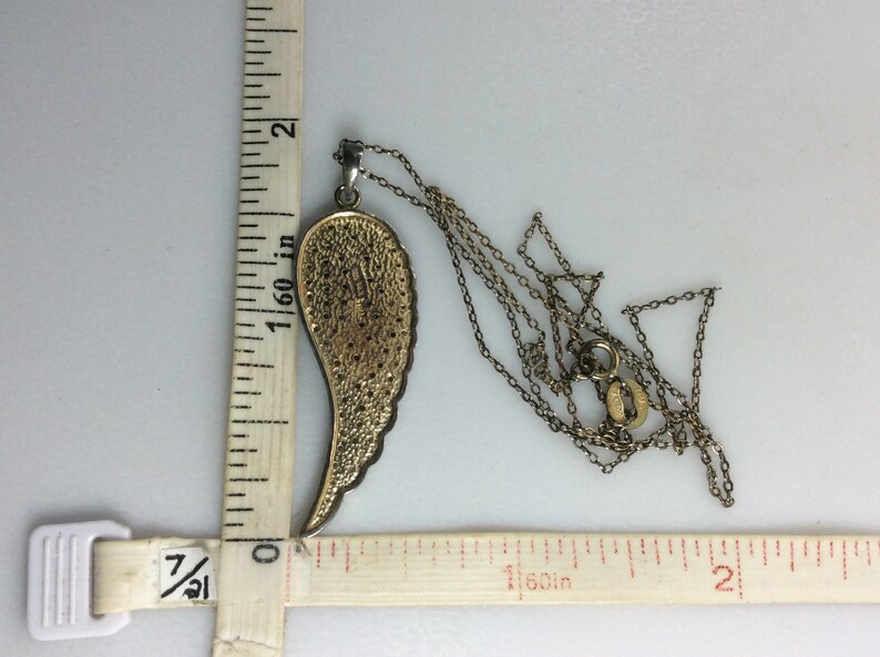 K&ouml;nnte beinhalten: Eine goldfarbene Engelsfl&uuml;gel-Anh&auml;ngerkette mit einer Kette, die etwa 3,8 cm lang ist. Der Anh&auml;nger ist etwa 3,8 cm lang und hat ein detailliertes Design.