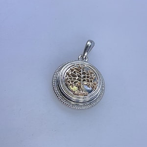 May include: Silver-tone pendant with a gold-colored tree of life design. The round pendant has a textured border and a small loop for a chain. The tree design is detailed and centered within the pendant.