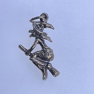 May include: A silver-toned charm depicting a witch riding a broom. The witch is wearing a pointed hat and flowing robes. The broom has a detailed handle and bristles. The charm is suspended from a small loop.