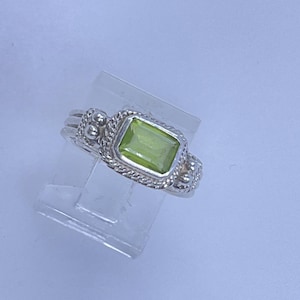 May include: A silver ring featuring a rectangular, light green gemstone. The ring has a detailed design with a braided border around the stone and decorative accents on the band. The ring is displayed on a clear stand.