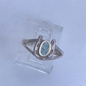 May include: A silver ring featuring a horseshoe design with a turquoise-colored stone in the center. The ring is displayed on a clear plastic stand against a light background. The ring's band is simple and thin.
