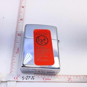 Puede incluir: Un encendedor Zippo cromado con una etiqueta de advertencia roja. La etiqueta presenta un gr&aacute;fico y el texto "KEEP AWAY FROM CHILDREN". Una cinta m&eacute;trica muestra el tama&ntilde;o aproximado del encendedor.