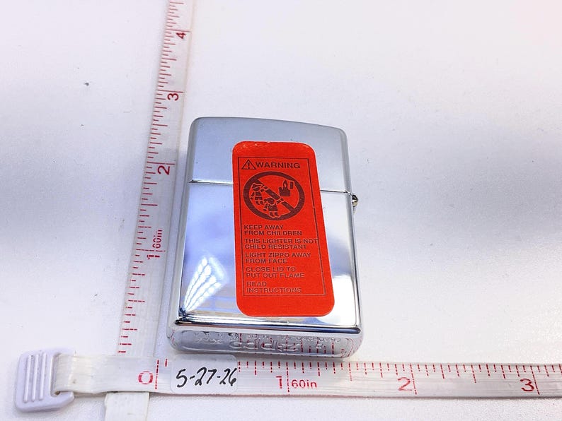 Puede incluir: Un encendedor Zippo cromado con una etiqueta de advertencia roja. La etiqueta presenta un gr&aacute;fico y el texto "KEEP AWAY FROM CHILDREN". Una cinta m&eacute;trica muestra el tama&ntilde;o aproximado del encendedor.
