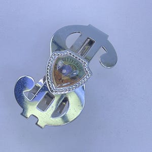 May include: A silver-colored money clip in the shape of a dollar sign. The clip features a shield-shaped emblem with the word "Gettysburg PA" on it. The emblem is surrounded by a decorative border.
