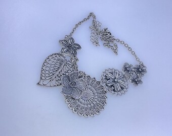 May include: Silver-toned statement necklace featuring intricate floral and leaf designs. The necklace has a chain with multiple decorative elements, including openwork leaves and detailed flower pendants. A unique piece of jewelry.