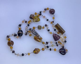 May include: A long necklace featuring a variety of glass beads in shades of brown, amber, and purple. The beads are strung on a silver-colored chain, with a mix of round, oval, and cylindrical shapes. The necklace is displayed against a white background.