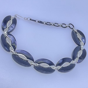 May include: A silver-toned bracelet featuring large, oval, dark blue links. The links are connected by smaller silver chains. The bracelet has a hook clasp. The jewelry is displayed on a light blue surface.