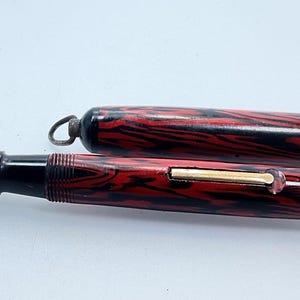 May include: A red and black patterned fountain pen with a gold trim and a matching cap. The pen has a black nib and a black and red striped barrel.