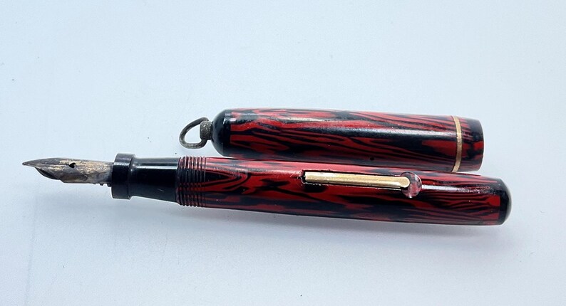 May include: A red and black patterned fountain pen with a gold trim and a matching cap. The pen has a black nib and a black and red striped barrel.