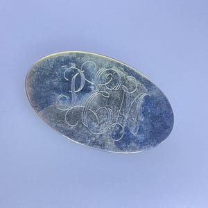May include: Oval silver-toned object with engraved script initials "PM" on a light blue background. The object has a slightly textured surface and a polished edge, reflecting light.