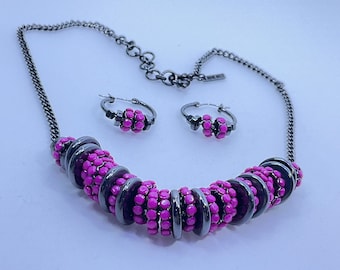 May include: A necklace and matching hoop earrings set. The necklace features a dark metal chain with a series of dark metal rings and bright pink beads. The earrings have a similar design, with dark metal hoops and pink beads.
