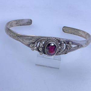 May include: A silver cuff bracelet featuring an oval-shaped deep red gemstone at the center. The metal band displays intricate wirework, small silver spheres, and leaf-like engravings surrounding the stone, with an open back design.