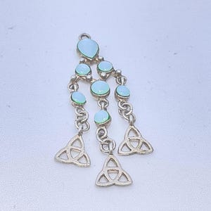 May include: A silver-toned pendant featuring a vertical arrangement of six round and teardrop-shaped light blue cabochons. The structure includes three dangling metal triquetra symbols connected by silver-toned chain links and small metal beads.