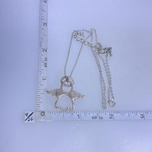 May include: A silver-colored pendant necklace featuring an angel design. The necklace is shown with a measuring tape, and the pendant is approximately 1.5 inches wide. The necklace has a delicate chain and a lobster claw clasp.