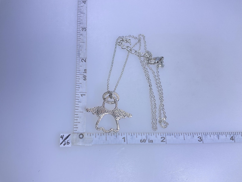 May include: A silver-colored pendant necklace featuring an angel design. The necklace is shown with a measuring tape, and the pendant is approximately 1.5 inches wide. The necklace has a delicate chain and a lobster claw clasp.