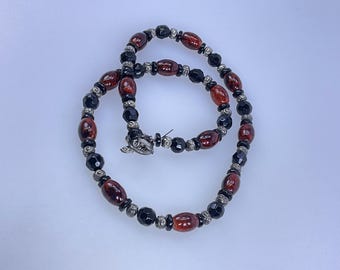 May include: A necklace with alternating dark red oval beads and black faceted beads. The necklace has silver-toned metal spacers and a decorative clasp. The jewellery is displayed on a white background.