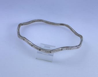 May include: A silver-colored, wavy bangle bracelet. The bracelet has an irregular shape and is decorated with small, circular patterns. The bracelet is displayed on a clear stand against a light background.