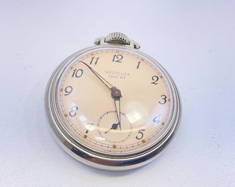 Op de afbeelding: Een vintage Westclox Pocket Ben zakhorloge met een zilverkleurige kast en een crèmekleurige wijzerplaat. Het horloge heeft zwarte cijfers, een kleine secondenwijzer en het Westclox-logo. Het horloge heeft een diameter van ongeveer 5 cm.