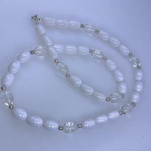 May include: A white beaded necklace with oval-shaped beads and clear, oval-shaped accent beads. The necklace has a silver clasp and silver accents between the beads. The beads are strung together to form a necklace.