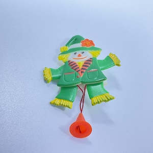 May include: A vintage plastic pull-string toy in the shape of a scarecrow. The scarecrow has a green jacket and pants, a yellow straw-like fringe, and a green hat with an orange flower. The pull string ends with an orange cone.