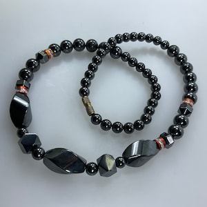 May include: A black hematite necklace and bracelet set with orange and silver accents. The necklace has larger, faceted hematite beads and the bracelet has smaller, round beads.