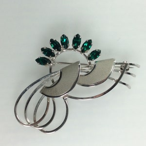 May include: A silver tone brooch with a design of overlapping circles and a fan shape. The brooch is accented with a row of emerald green rhinestones.