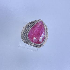 May include: A silver ring featuring a large, pear-shaped, faceted ruby gemstone. The ring's band has a detailed, textured design with small, circular accents. The ruby is a vibrant shade of pink.