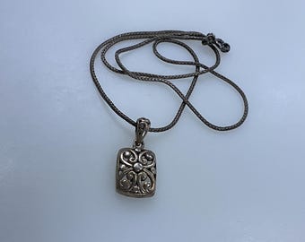 May include: A silver necklace with a square pendant featuring a detailed floral design. The pendant is attached to a thin, braided cord.