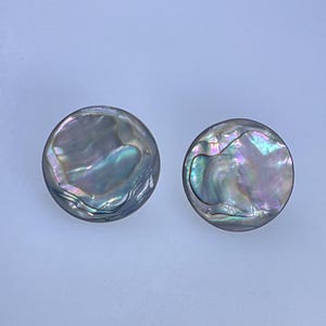 May include: Two round, iridescent abalone shell buttons. The buttons have a pearlescent sheen with colors of blue, green, and purple. The buttons are set against a light blue background.