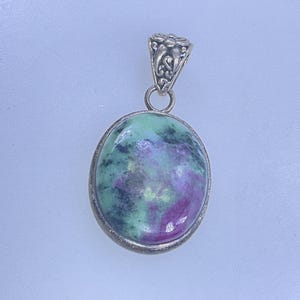 May include: Oval pendant with a silver-colored setting. The pendant features a polished stone with a mix of green and purple hues. The bail is decorated with a floral design.