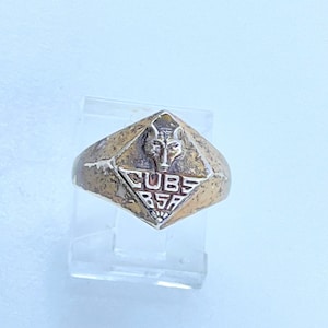 May include: A gold-toned ring with a diamond-shaped face featuring a wolf head and the words "CUBS BSA".