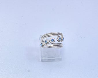 May include: A silver-toned open-style ring featuring multiple swirling bands that converge at the ends. The design is accented with five small, round, bright blue stones embedded within the metal swirls, creating a wave-like appearance.