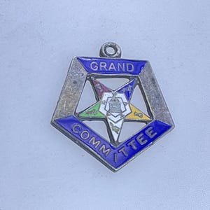 May include: A silver-toned pendant with a blue border and the words "GRAND COMMITTEE". The pendant features a star with colorful enamel details. The pendant has a small loop at the top for attachment.