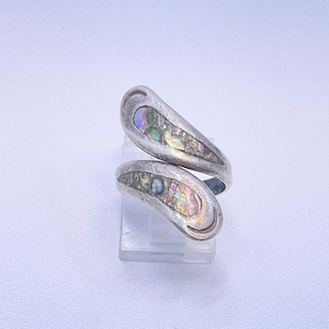 May include: A bypass style ring crafted from silver-toned metal, featuring two tapered ends that overlap. Each end is inlaid with iridescent abalone shell segments displaying a spectrum of green, blue, and pink hues against a polished surface.