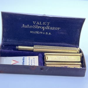 May include: An open, vintage, dark blue velvet-lined case containing a gold-colored Valet AutoStrop Razor, a razor blade, and a handle. The case is marked "MADE IN U.S.A."