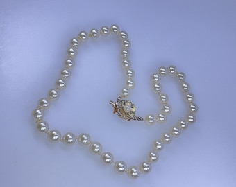 May include: Cream-colored pearl necklace with a gold-tone clasp. The necklace features round, evenly spaced pearls. The clasp has a floral design with a small pearl in the center. The necklace is displayed on a white surface.