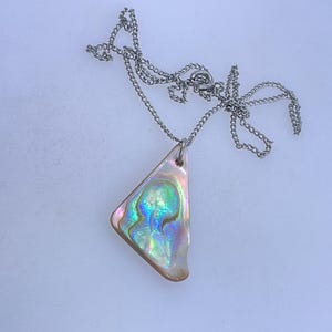May include: Abalone shell pendant necklace. The pendant is a triangular shape with iridescent colors of blue, green, and pink. The necklace chain is silver-colored and delicate. The necklace is displayed on a white surface.