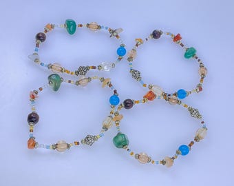 May include: Four beaded bracelets with a variety of colorful beads, including green, blue, orange, and brown. The bracelets have a mix of shapes and sizes, with some beads appearing to be glass or crystal. The bracelets are arranged on a white surface.