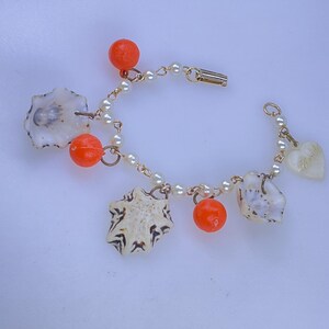 May include: A delicate bracelet featuring a gold chain, small pearls, and seashell charms. The bracelet is adorned with orange beads and a heart-shaped charm. The seashells have a unique pattern, and the bracelet is secured with a gold clasp.