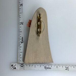 May include: A wooden brooch in the shape of a tall, narrow, pointed structure. The brooch is painted a light beige color and has a gold-toned pin on the back.