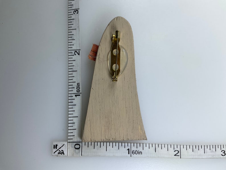 May include: A wooden brooch in the shape of a tall, narrow, pointed structure. The brooch is painted a light beige color and has a gold-toned pin on the back.