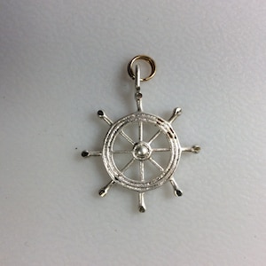 May include: Silver ship's wheel pendant with a gold jump ring. The wheel has eight spokes and a small round center.
